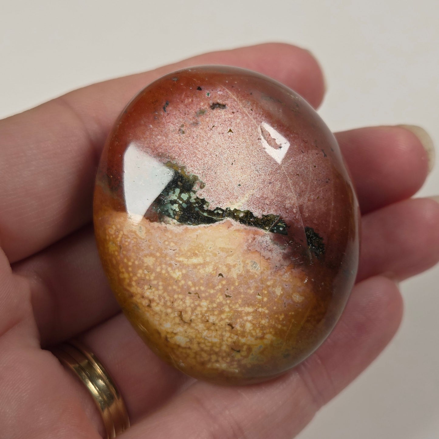 A hand holding a polished oval stone with a mottled pattern of reddish-brown, gold, and dark green.