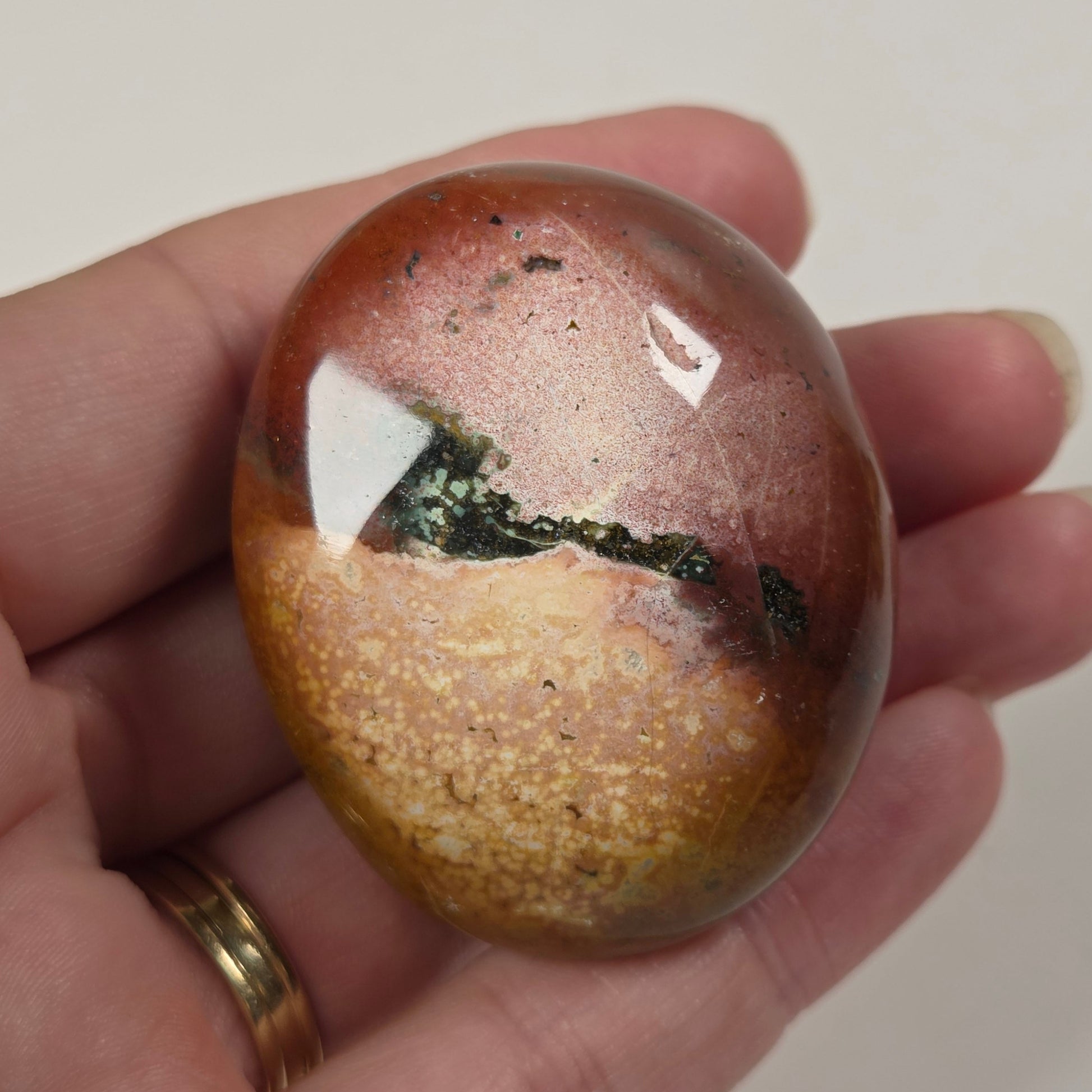 A hand holding a polished oval stone with a mottled pattern of reddish-brown, gold, and dark green.