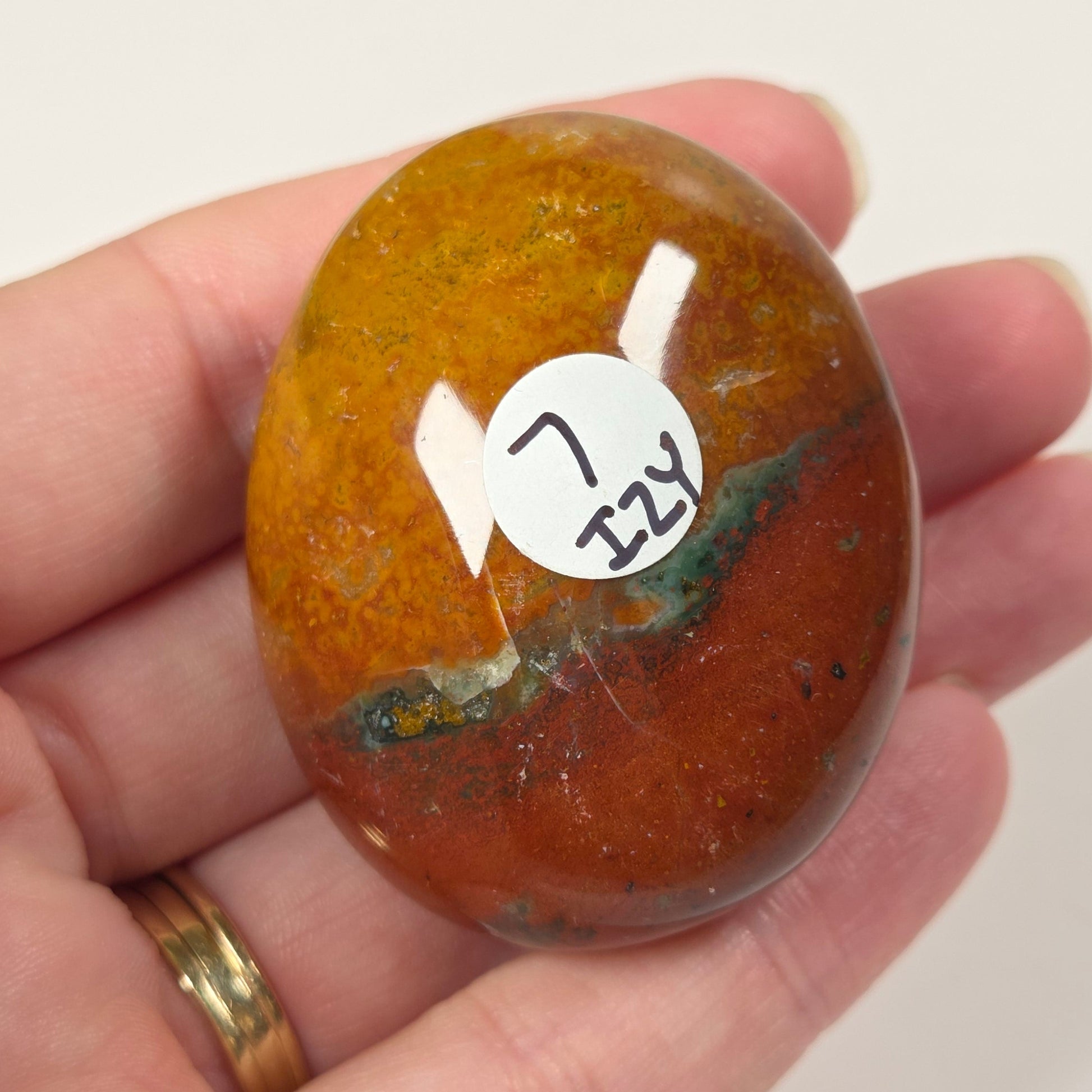 A hand holding a polished oval gemstone with a mottled orange, brown, and green pattern, featuring a white sticker with handwritten text.