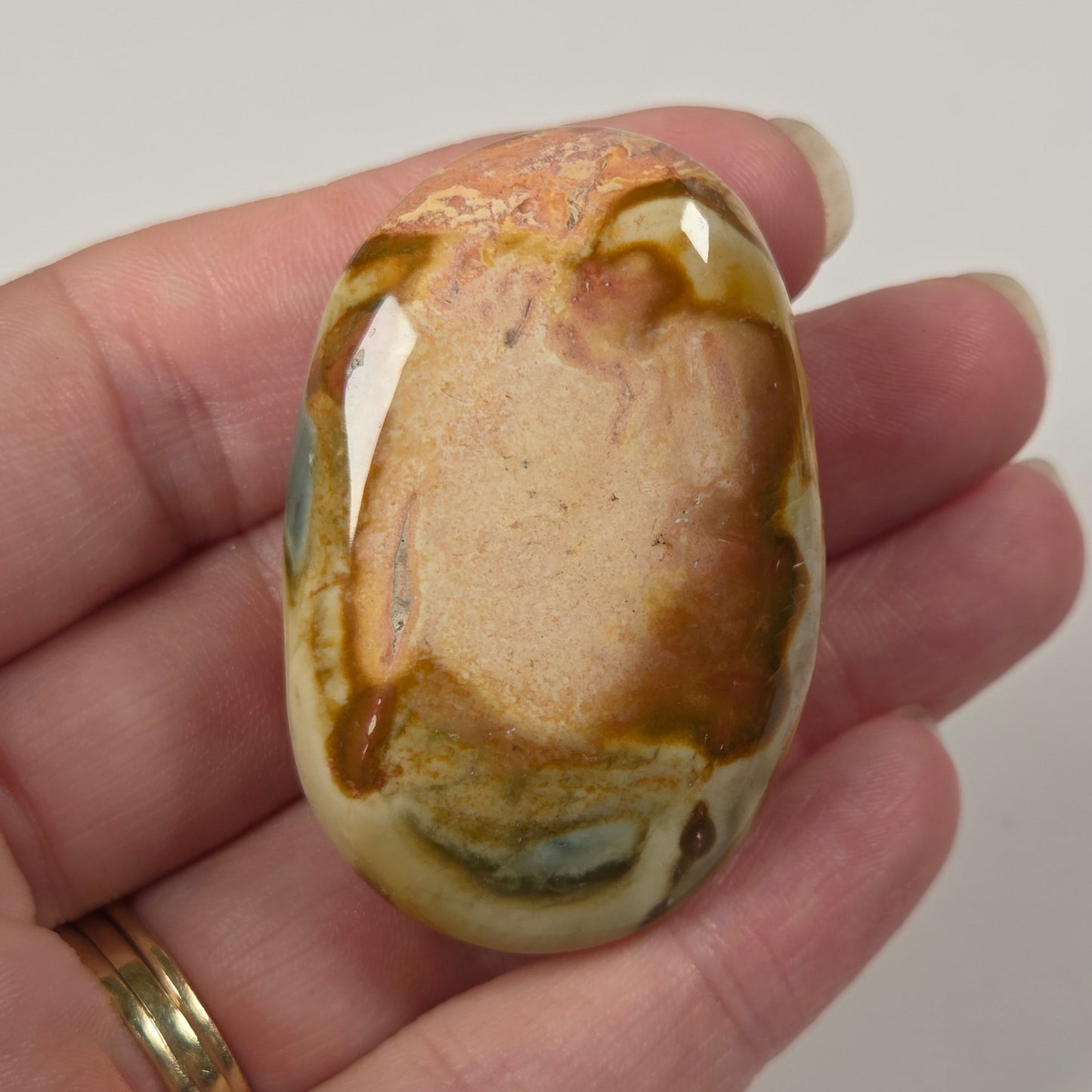 A polished oval stone with swirling tan, brown, cream, and muted green patterns, held in a person's hand against a plain light background.