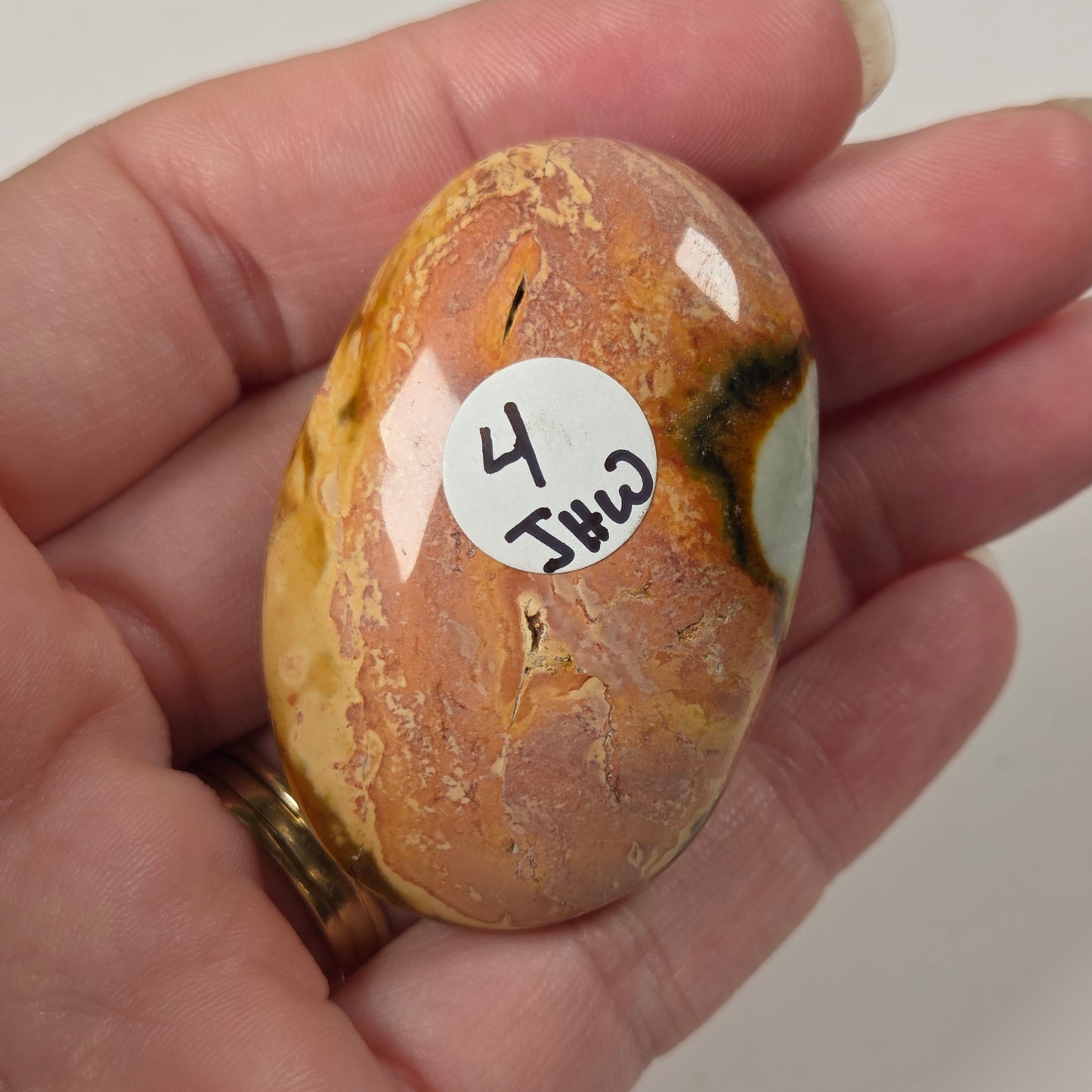 A hand holding a polished, oval-shaped brown and orange stone with natural marbling and a small white sticker labeled '4 Jhu 2'.