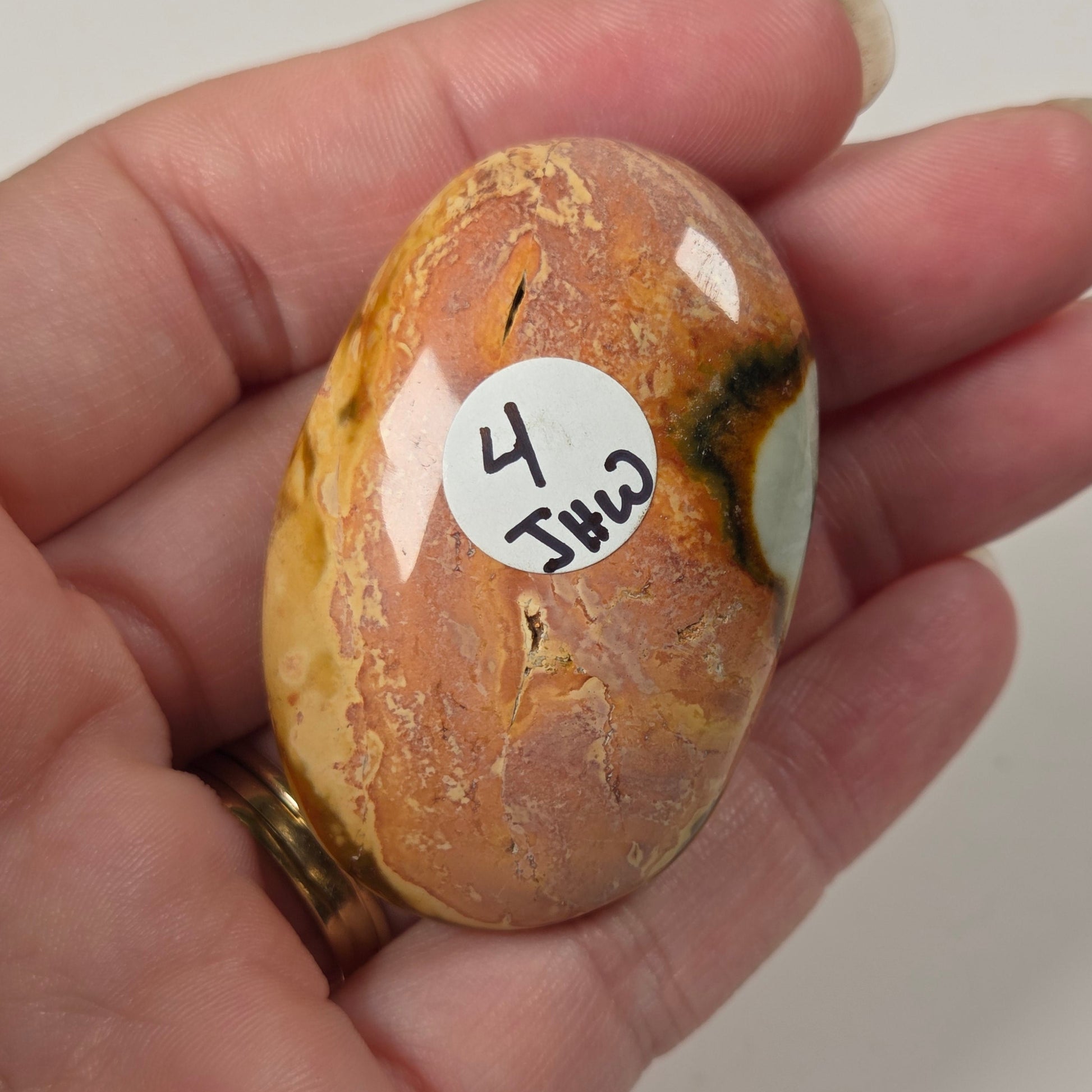 A hand holding a polished, oval-shaped brown and orange stone with natural marbling and a small white sticker labeled '4 Jhu 2'.