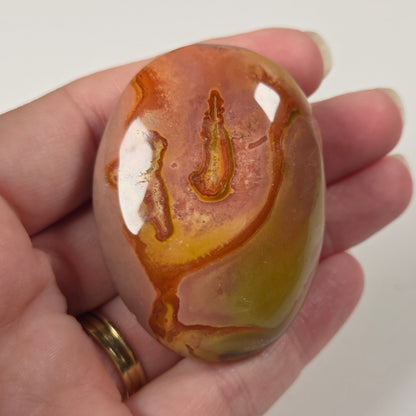 A hand holds a polished oval stone with swirling orange, yellow, and green agate-like patterns against a plain light background.