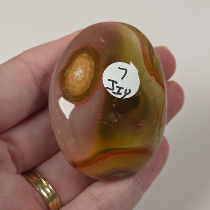 A hand holds a polished oval agate stone with swirling bands of brown, amber, and cream. A small white sticker with handwritten text is attached to the stone.