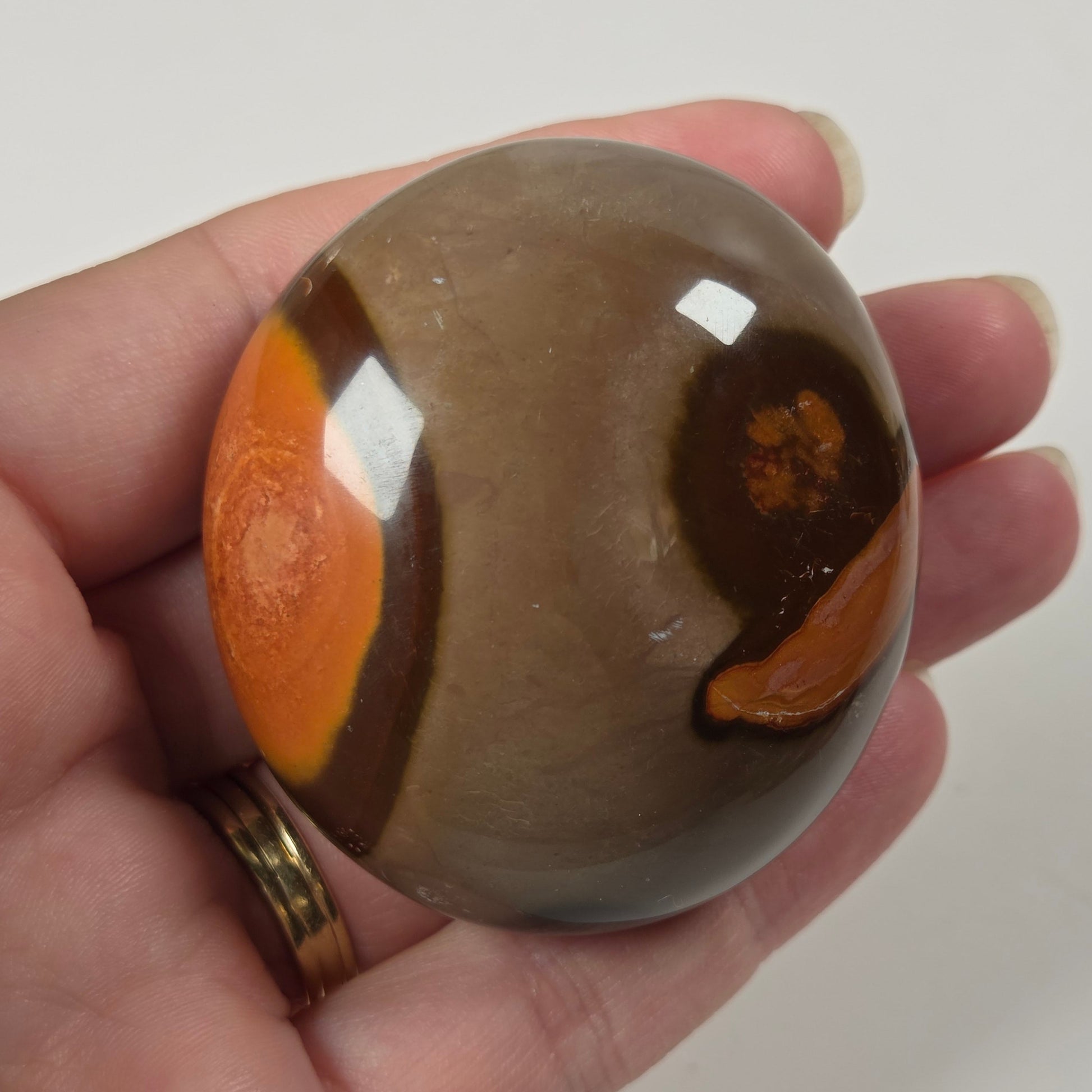 A hand holding a polished oval stone with a brown and gray body and bright orange inclusions, shown against a plain light background.