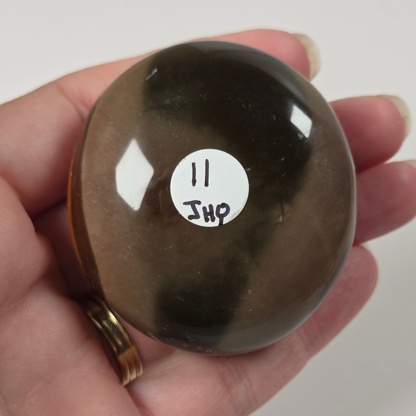A hand holds a smooth, polished oval brown stone with a white sticker labeled '11 JHP' on its surface, set against a plain light background.