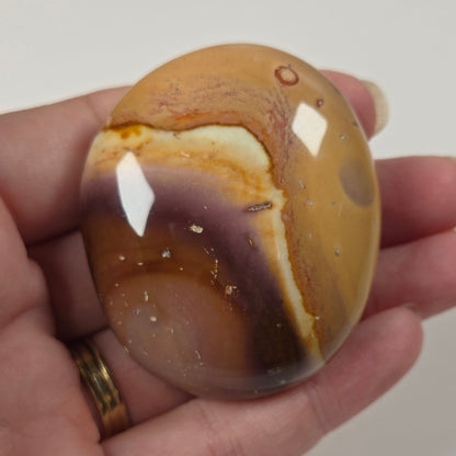 A hand holding a polished oval stone cabochon with a marbled pattern of brown, tan, cream, and dark purple, set against a plain light background.