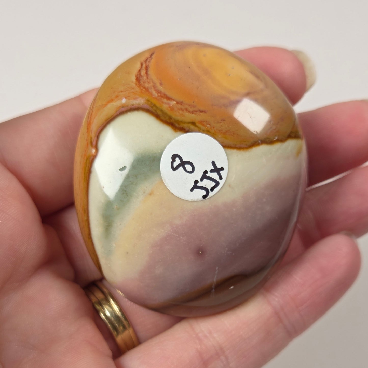 A hand holds a polished, rounded stone with marbled patterns of orange, brown, cream, and pale green. A small white sticker with the handwritten numbers '8 151' is attached to the stone.