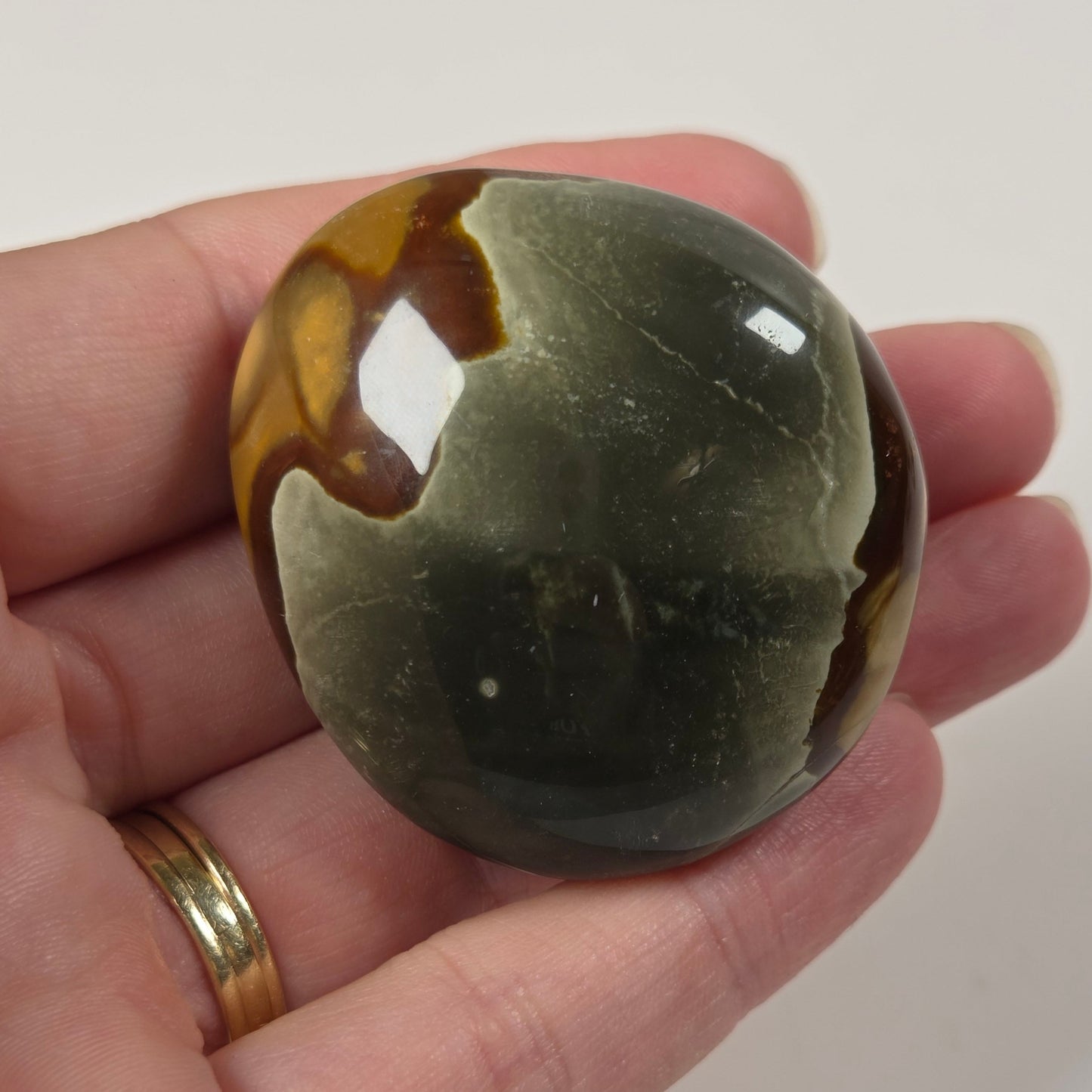 A polished stone ornament with a mottled pattern of olive green, dark gray, and brown, held in a person's hand against a plain light background.