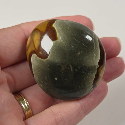A polished stone ornament with a mottled pattern of olive green, dark gray, and brown, held in a person's hand against a plain light background.