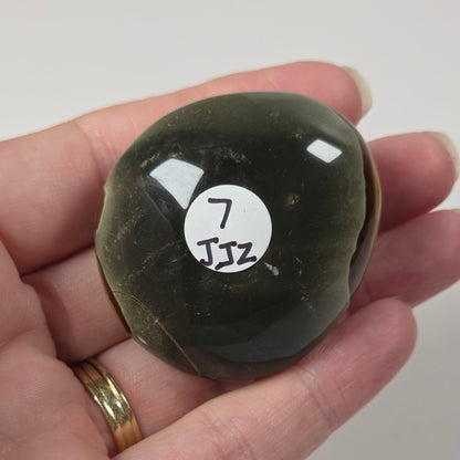 A hand holding a polished dark green stone with a white sticker labeled '7 JSZ' on its surface, set against a plain light background.