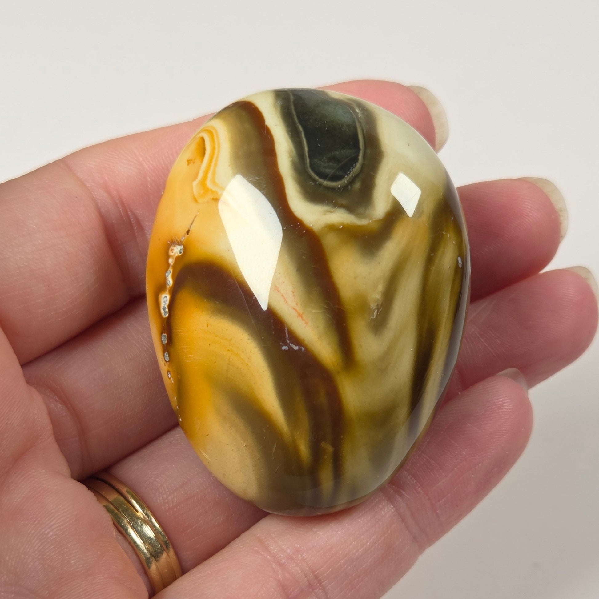 A polished oval agate stone with swirling bands of yellow, brown, black, and cream, held in a person's hand against a plain white background.