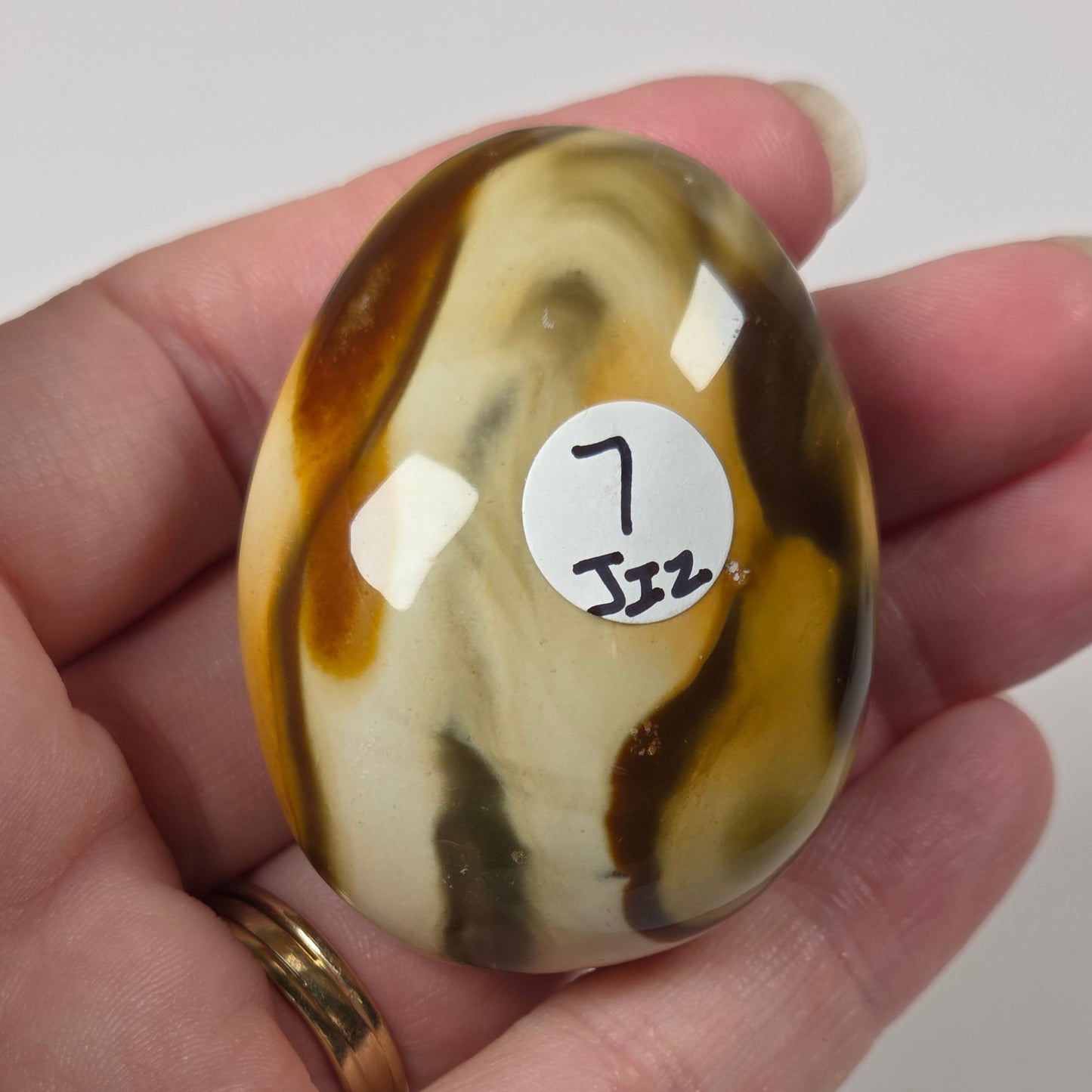 A hand holds a polished oval stone with marbled brown, cream, and dark gray patterns. A small white sticker on the stone reads '7 Jzz'.