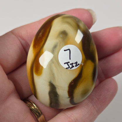 A hand holds a polished oval stone with marbled brown, cream, and dark gray patterns. A small white sticker on the stone reads '7 Jzz'.