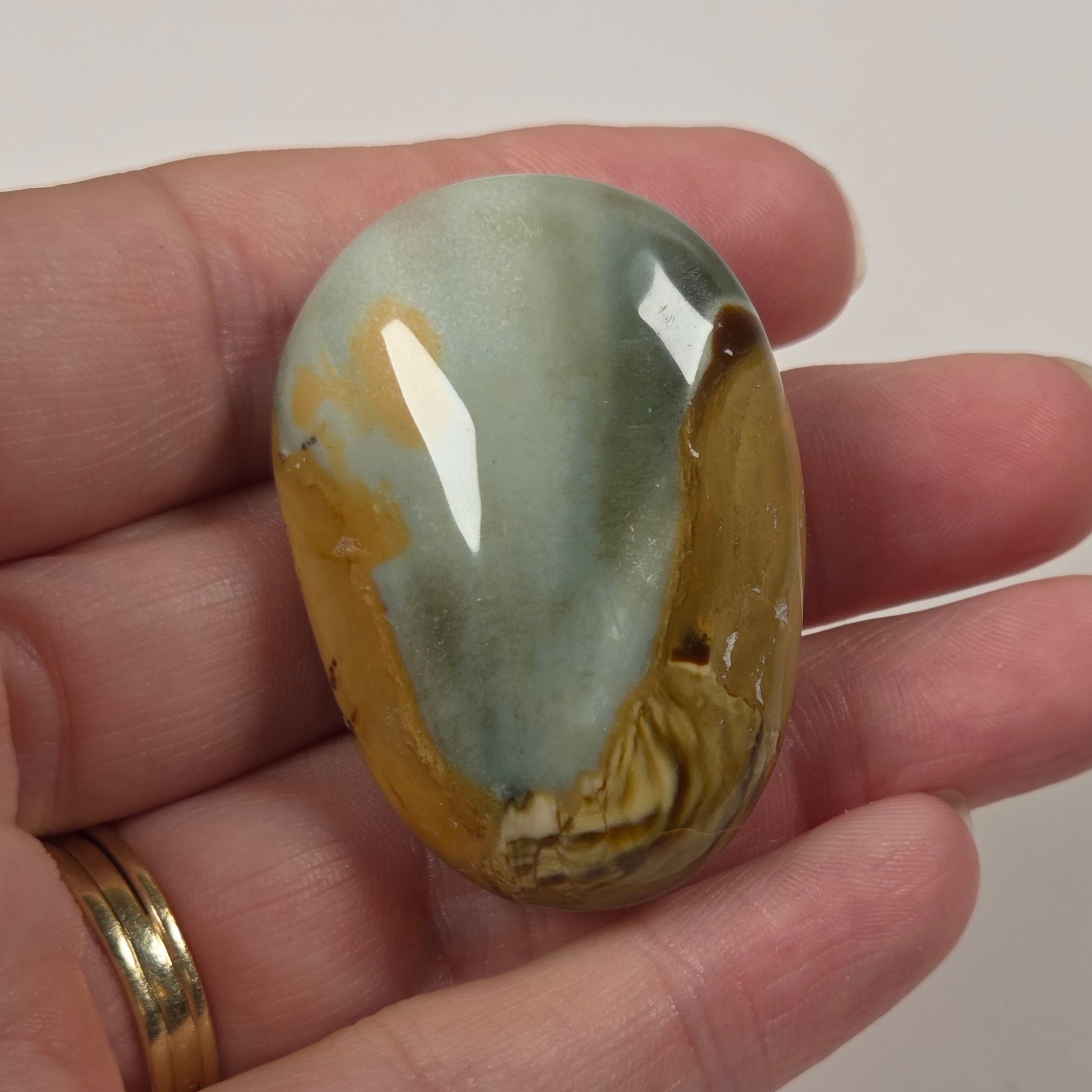 A hand holding a polished oval stone cabochon with a marbled pattern of pale blue-green, cream, and golden yellow, set against a plain light background.