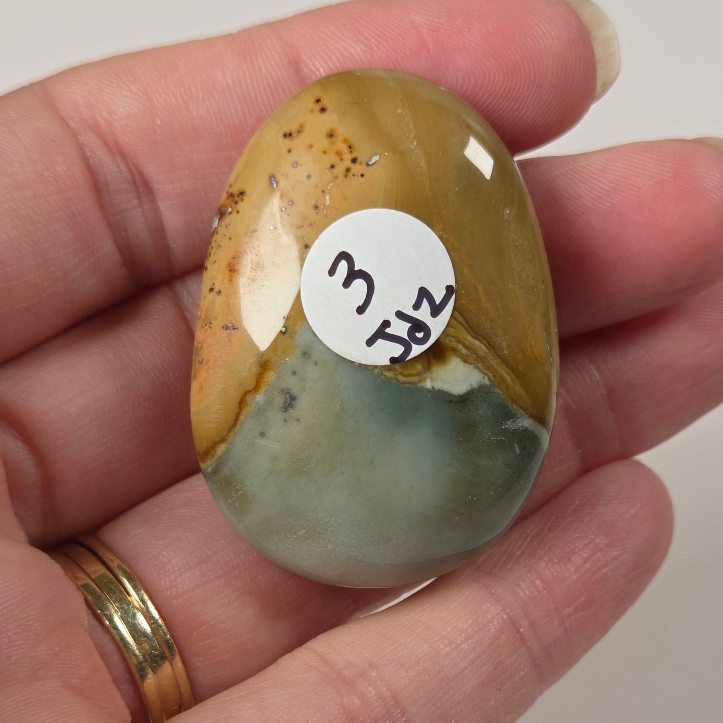 A hand holding a polished, tumbled stone cabochon with a white circular sticker reading '3 1/2'. The stone features a marbled pattern of golden yellow, brown, and pale green.