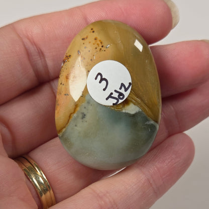 A hand holding a polished, tumbled stone cabochon with a white circular sticker reading '3 1/2'. The stone features a marbled pattern of golden yellow, brown, and pale green.