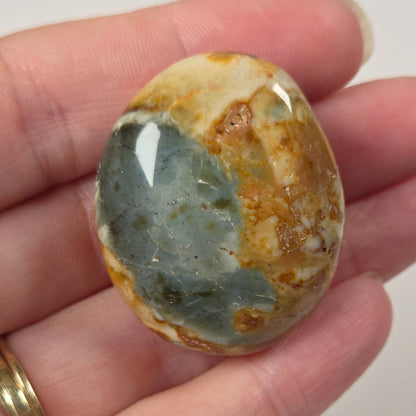 A polished oval stone with a mottled pattern of blue-green, tan, and brown, held in a person's hand against a plain light background.