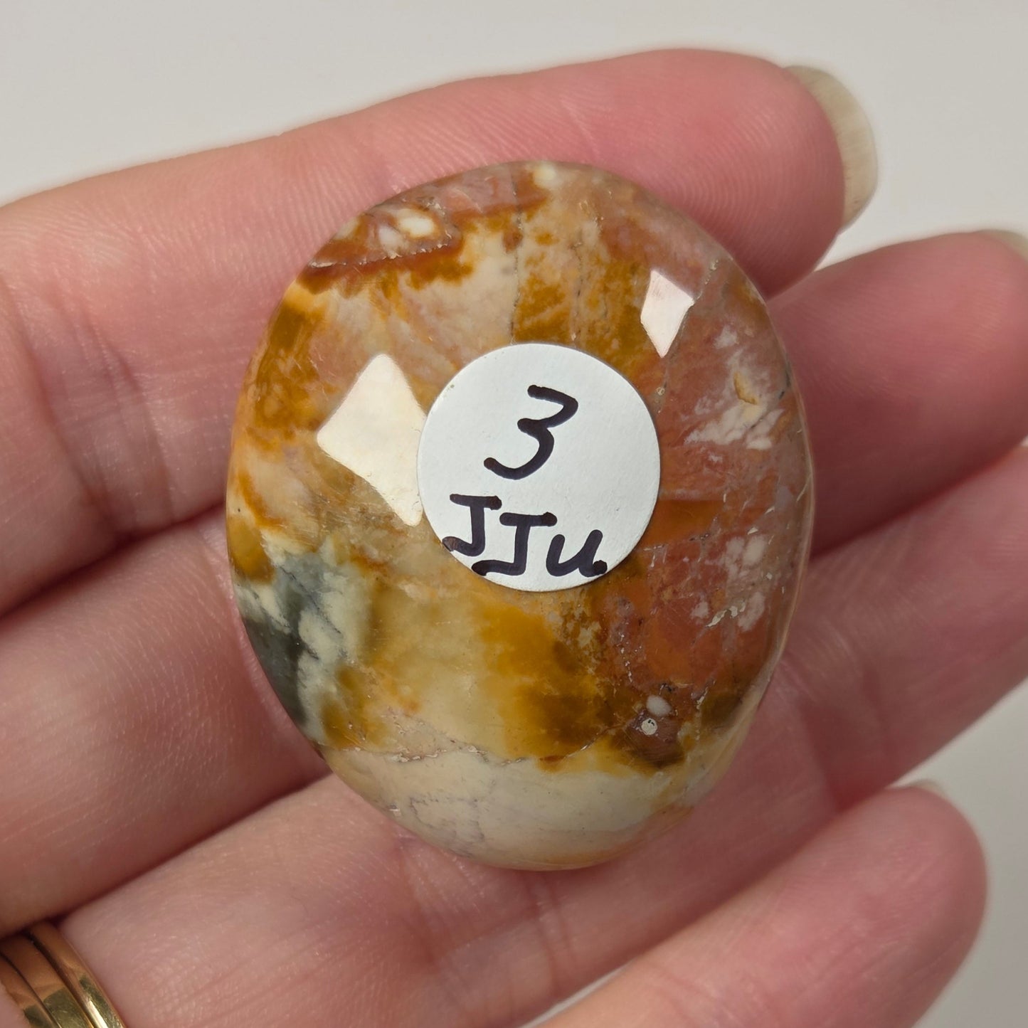 A hand holds a polished oval stone cabochon with marbled orange, brown, cream, and white patterns. A small white sticker with the handwritten text '3 JTu' is attached to the front of the stone.