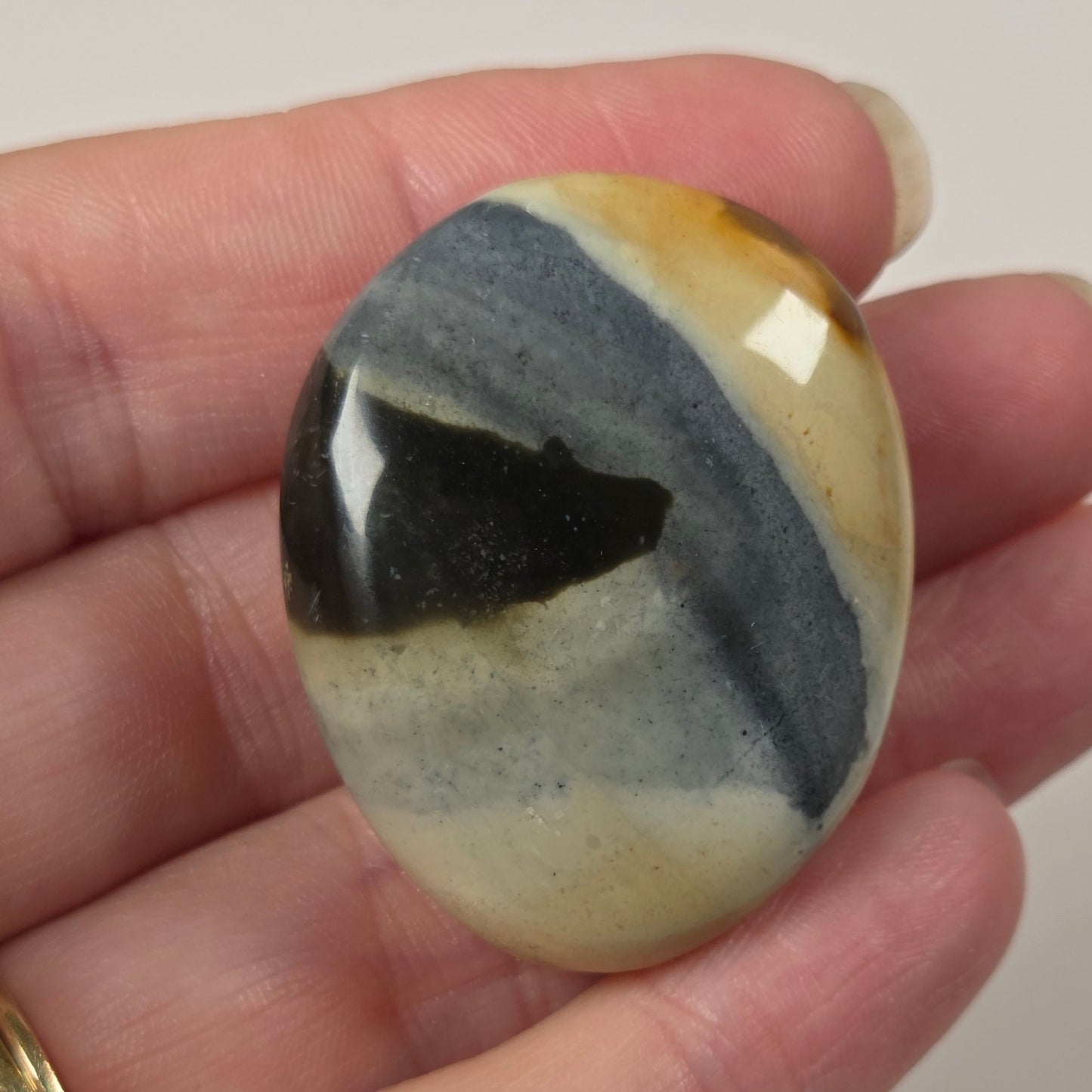 A hand holding a polished oval stone cabochon with a marbled pattern of black, slate blue, cream, and ochre yellow, set against a plain light background.