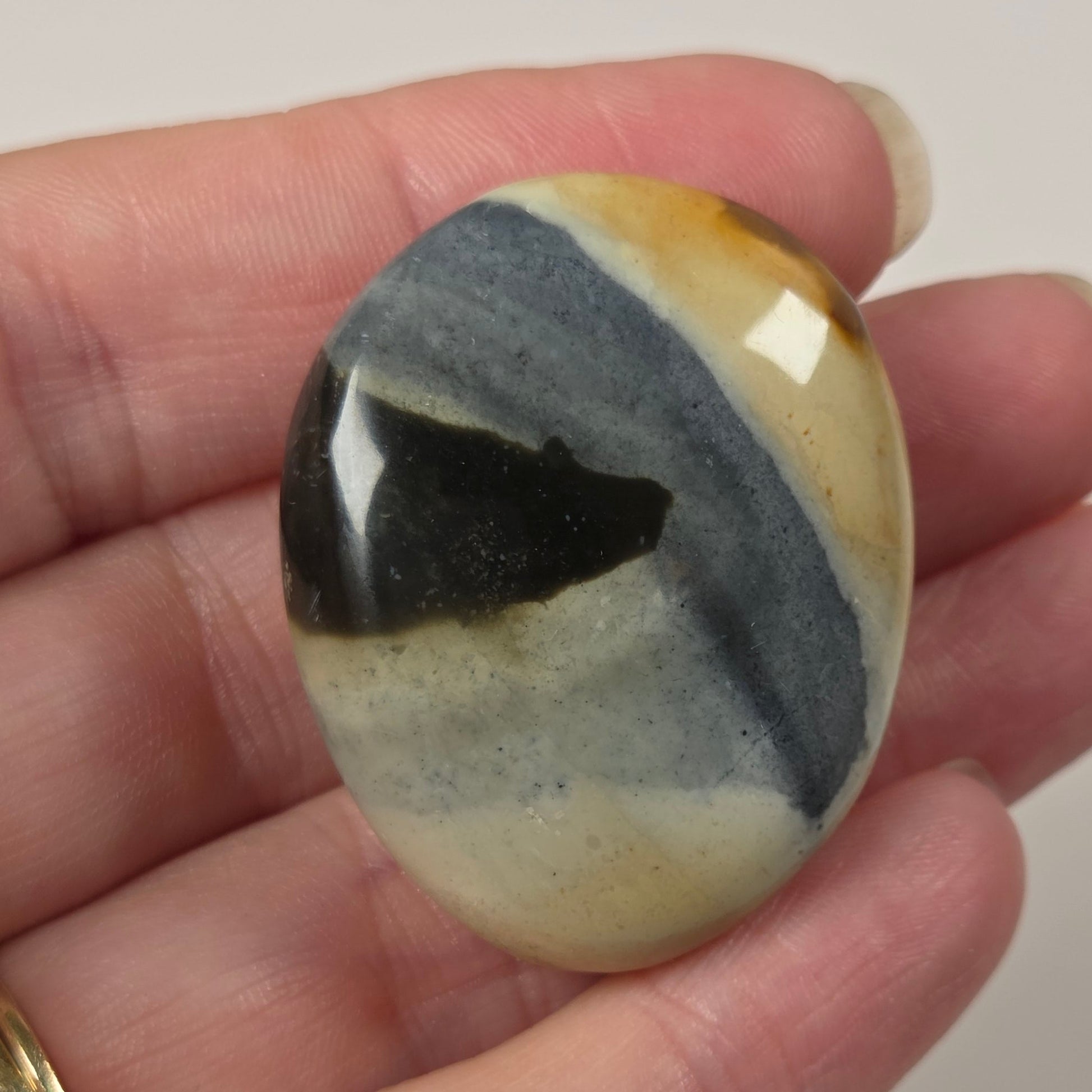 A hand holding a polished oval stone cabochon with a marbled pattern of black, slate blue, cream, and ochre yellow, set against a plain light background.