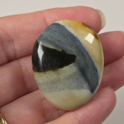 A hand holding a polished oval stone cabochon with a marbled pattern of black, slate blue, cream, and ochre yellow, set against a plain light background.