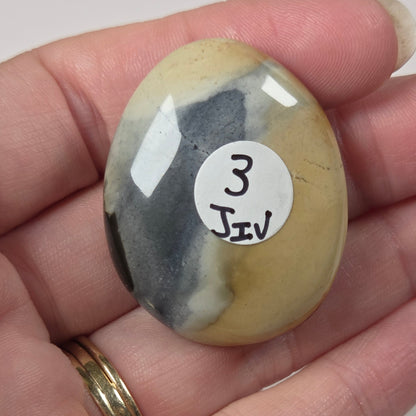 A hand holding a polished oval stone with a white label that reads '3 JIV'. The stone has a marbled pattern of yellow, beige, gray, and black, and the hand is wearing a gold ring against a plain light background.