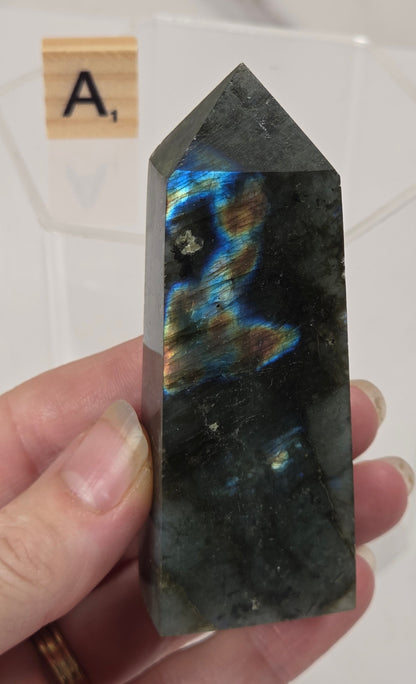 A hand holding a polished labradorite crystal point with vibrant blue, green, and gold iridescent flashes on a white background.