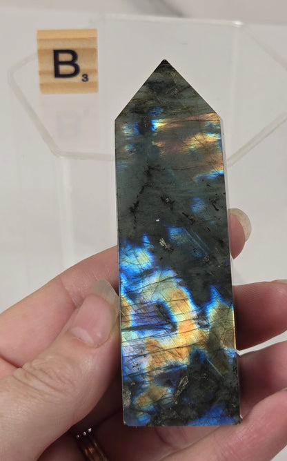 A hand holds a polished labradorite crystal point showing vibrant blue, gold, and green flashes. A small Scrabble tile with the letter 'B' is visible in the background against a white surface.