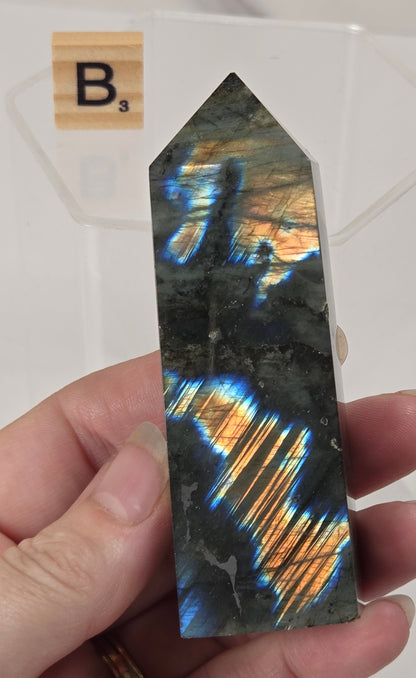 A hand holds a polished labradorite crystal point showing vibrant blue, orange, and gold flashes. A white background with a clear display block and a Scrabble tile marked B3 is visible behind it.