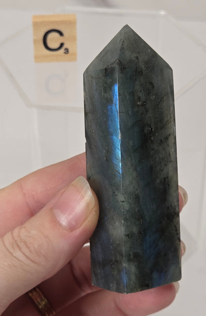 A hand holding a polished dark gray labradorite crystal point with a blue flash, with a clear display block and a Scrabble tile showing 'C3' in the background.