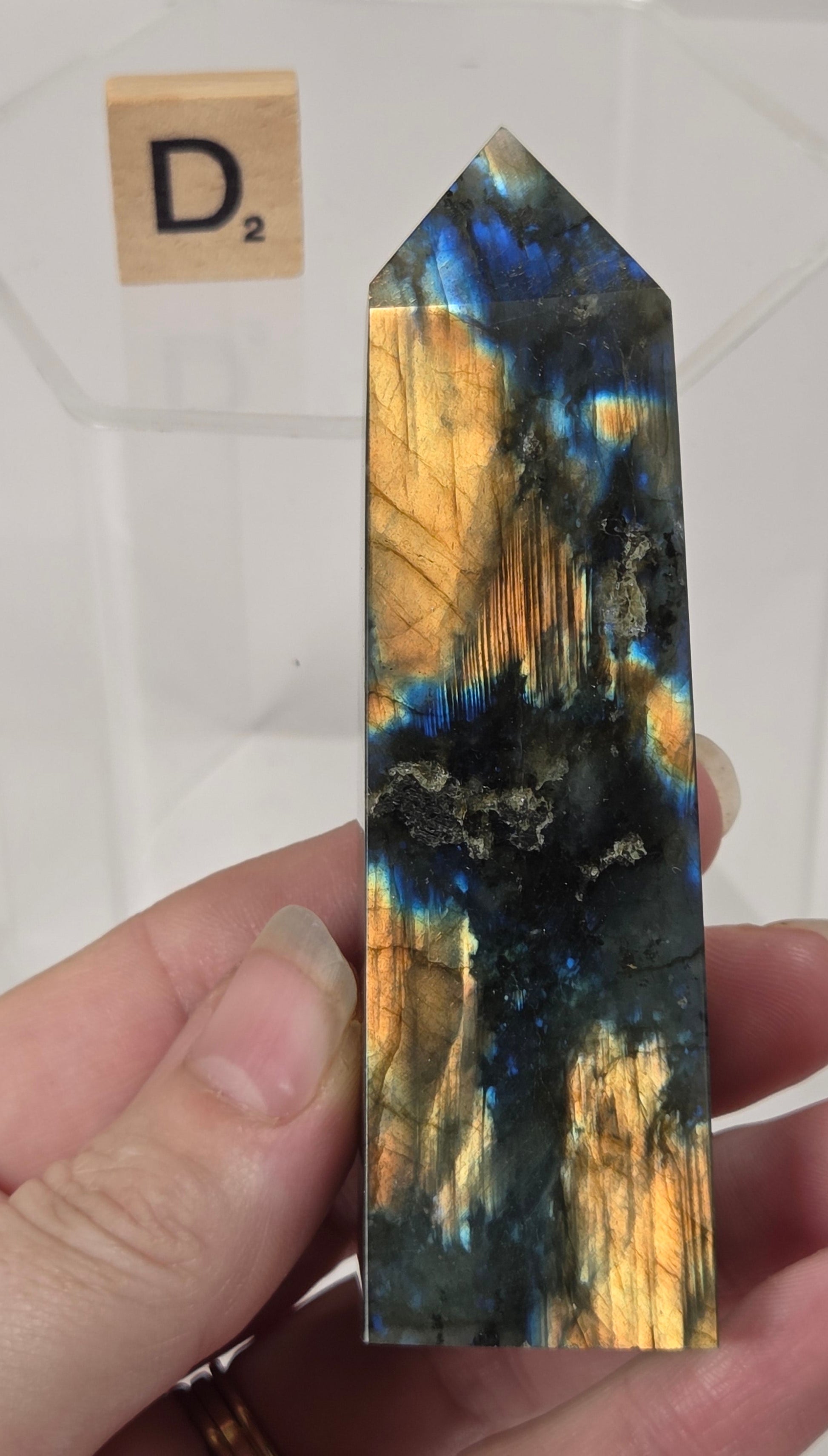 A hand holds a polished labradorite crystal tower pointing upward, showing a shimmering mix of gold, blue, and black iridescent colors. A clear display block and a Scrabble tile labeled 'D2' are blurred in the background.