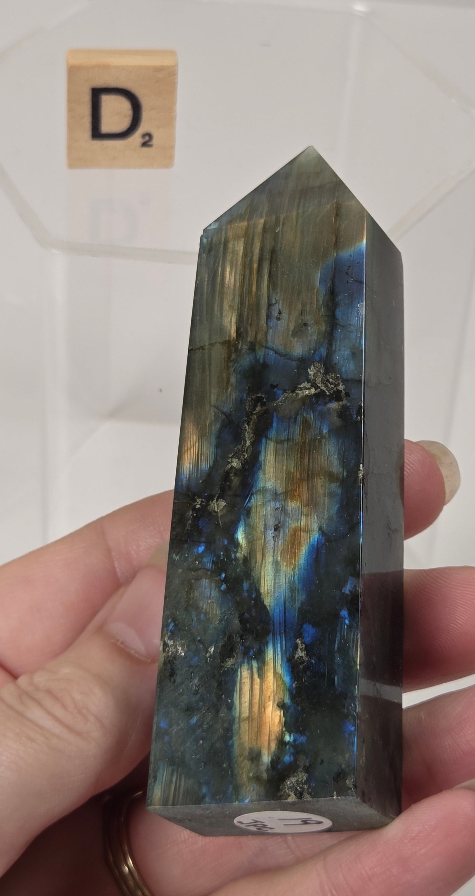 A hand holding a polished labradorite crystal point with natural blue, gold, and gray iridescence, with a clear display block and a Scrabble tile in the background.