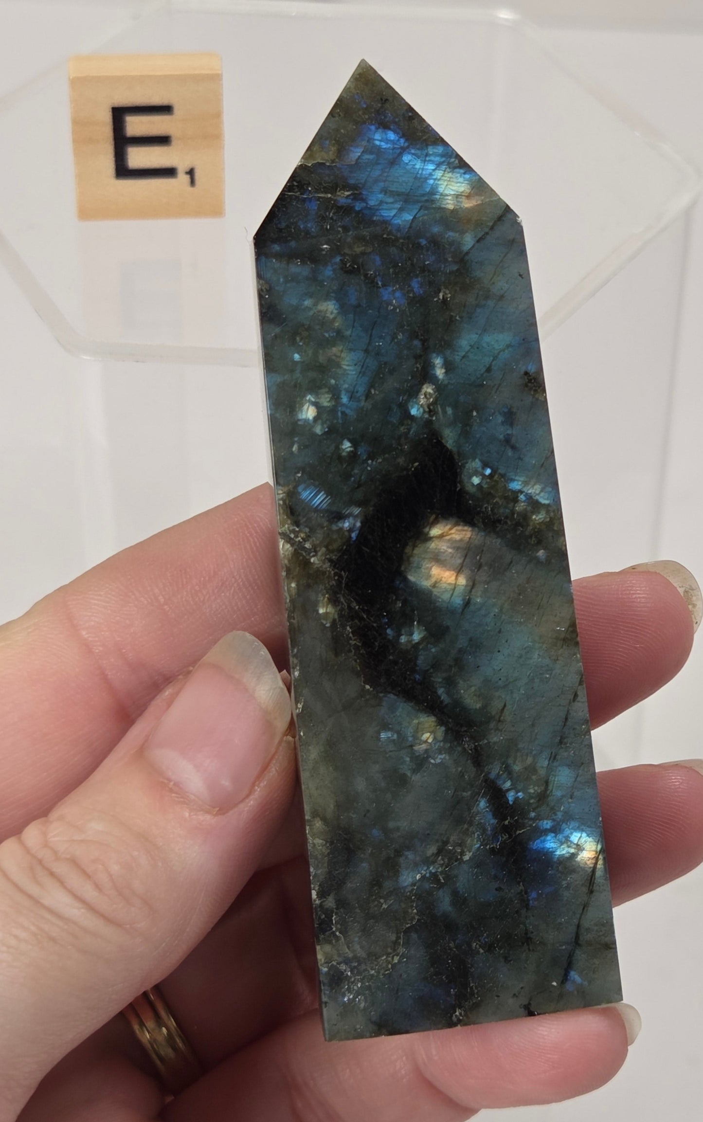 A hand holding a polished labradorite crystal point with shimmering blue, green, and black flashes, with a small wooden letter tile in the background.