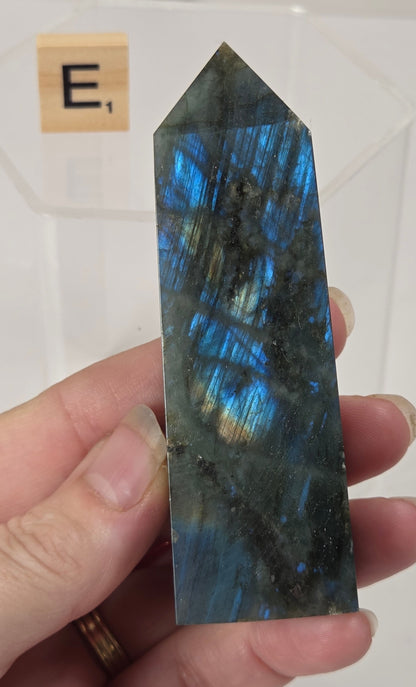 A hand holding a polished labradorite crystal point with shimmering blue, green, and gray flashes, with a small card labeled 'E' in the background.
