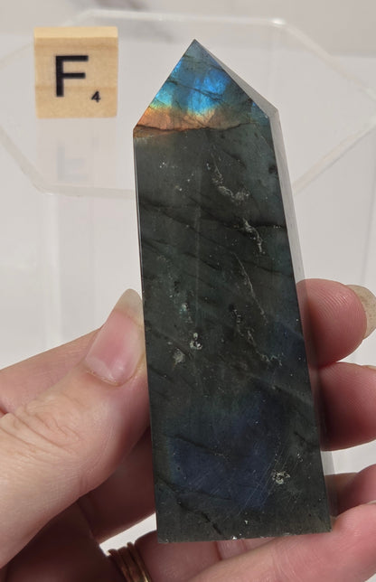 A hand holds a polished labradorite crystal point showing a blue and green iridescent sheen. A small Scrabble tile with the letter F is visible in the background on a white surface.