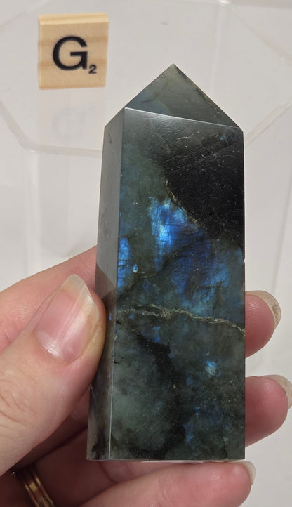 A hand holds a polished labradorite crystal tower showing vibrant blue flashes, with a small Scrabble tile in the background.