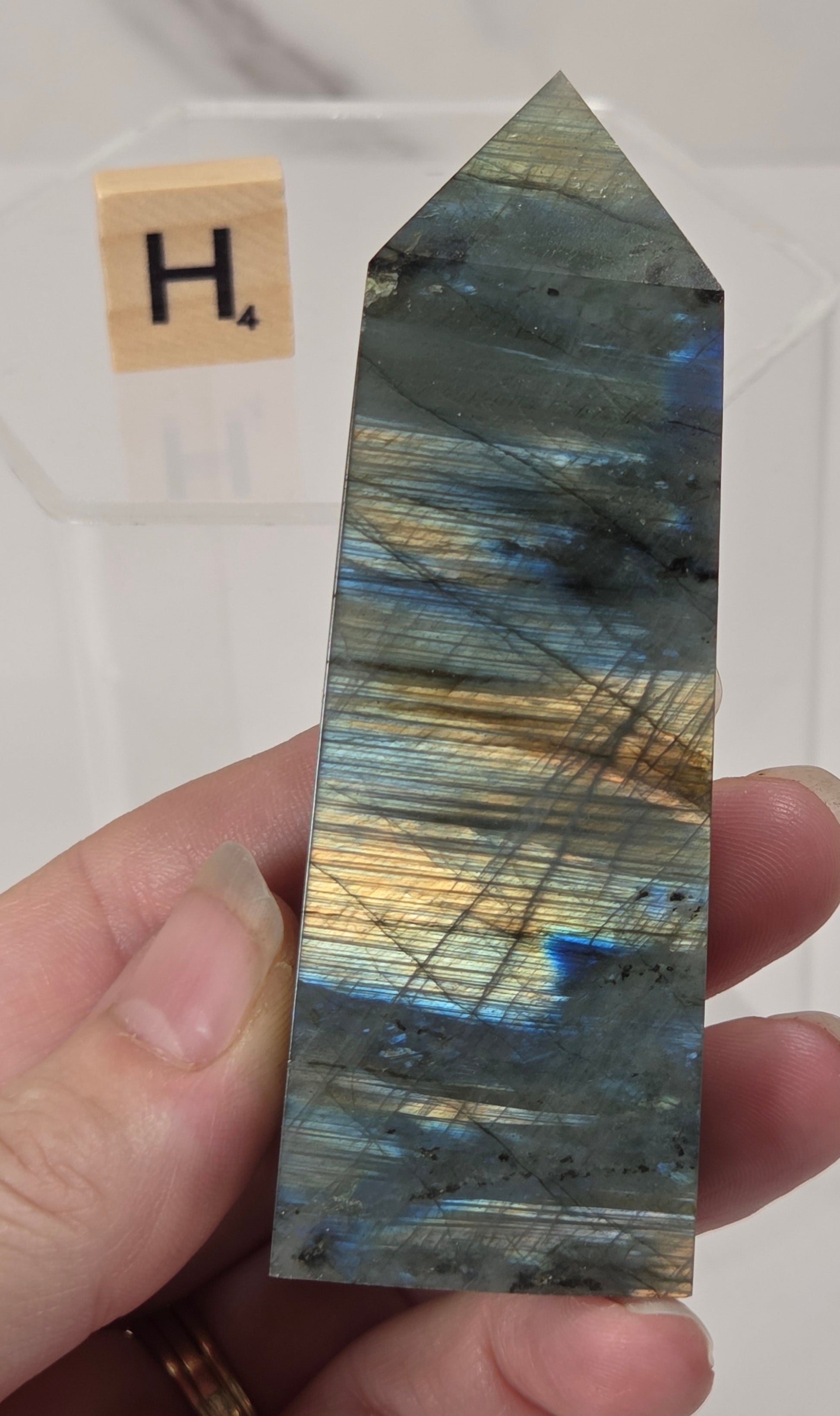 A hand holds a polished labradorite crystal point showing iridescent blue, gold, and gray flashes, with a small Scrabble tile in the background.