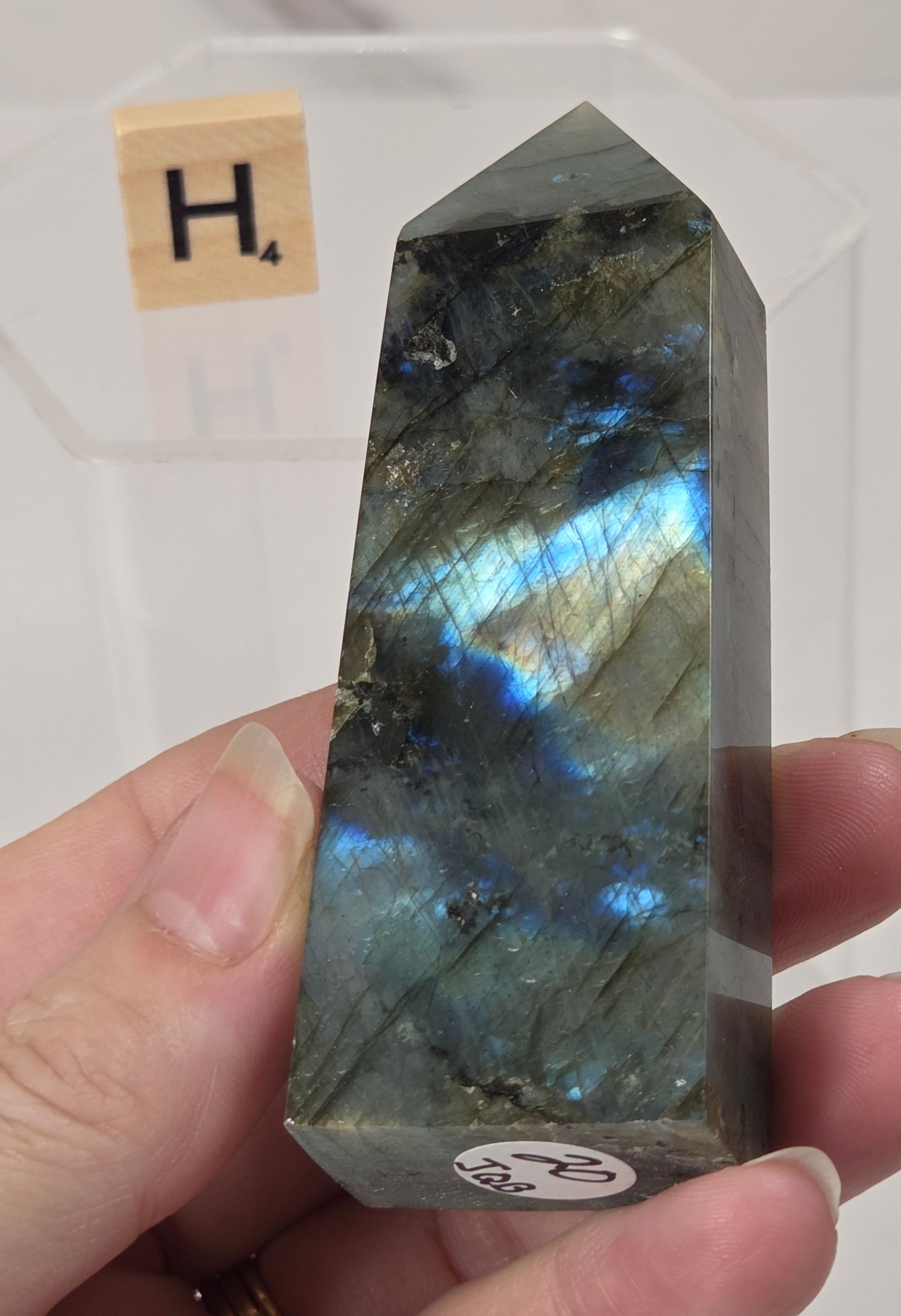 A hand holds a polished labradorite crystal point showing vibrant blue and green flashes, with a small white sticker at the base and a Scrabble tile in the background.