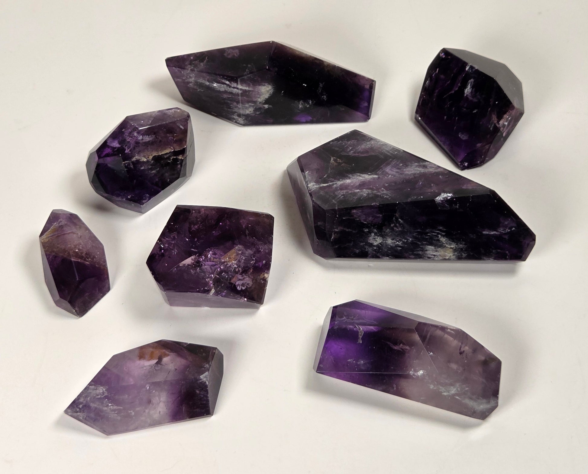 A set of eight raw amethyst crystal points and cuboids in various sizes, arranged on a plain white surface.