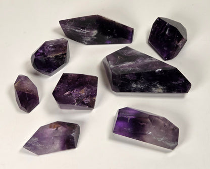 A set of eight raw amethyst crystal points and cuboids in various sizes, arranged on a plain white surface.