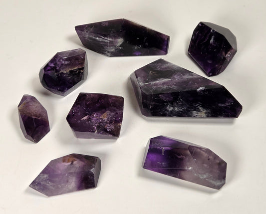 A set of eight raw amethyst crystal points and cuboids in various sizes, arranged on a plain white surface.