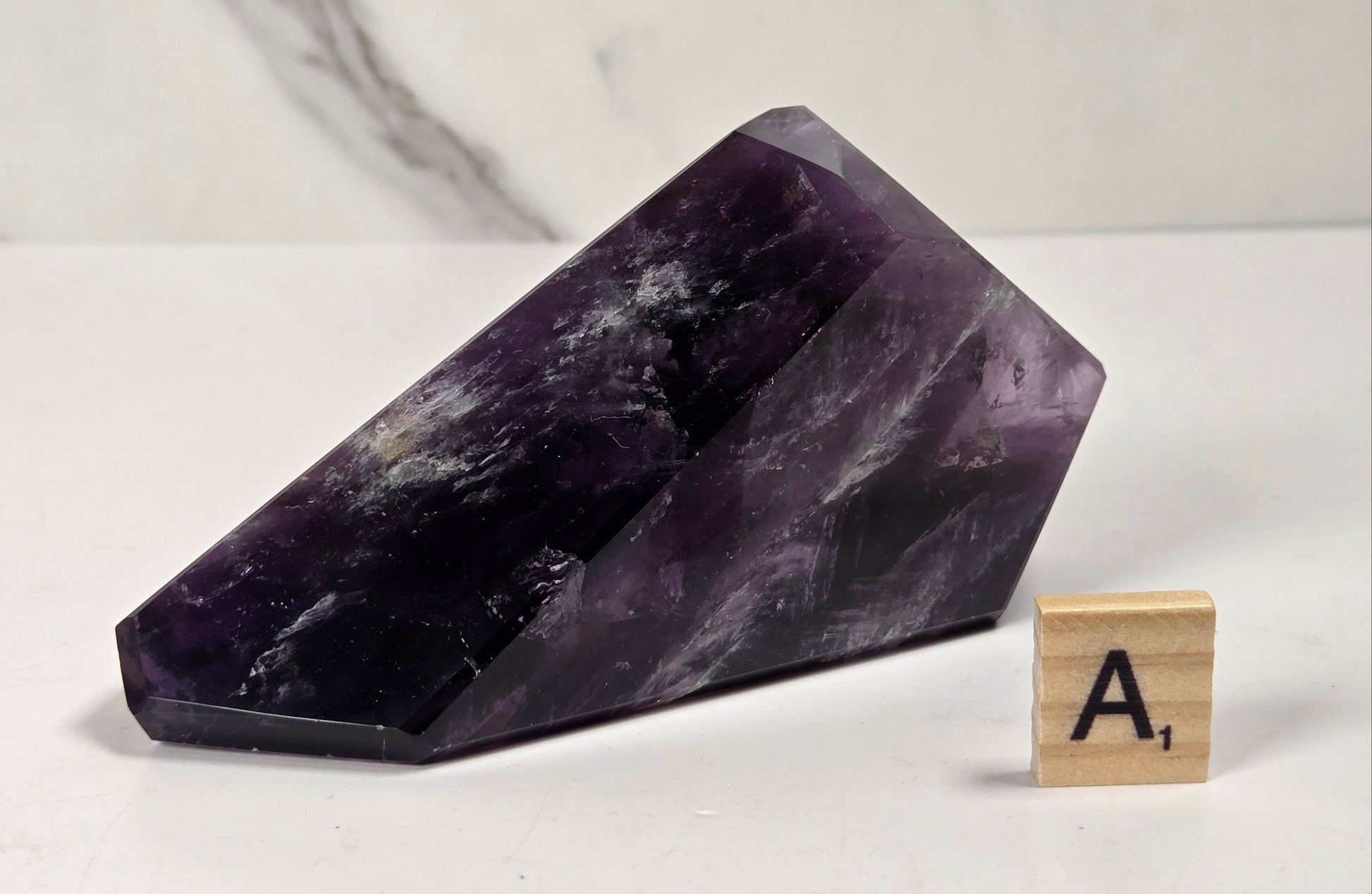 A polished purple amethyst crystal cluster with natural white and dark inclusions, displayed on a white surface next to a wooden Scrabble tile showing the letter A.