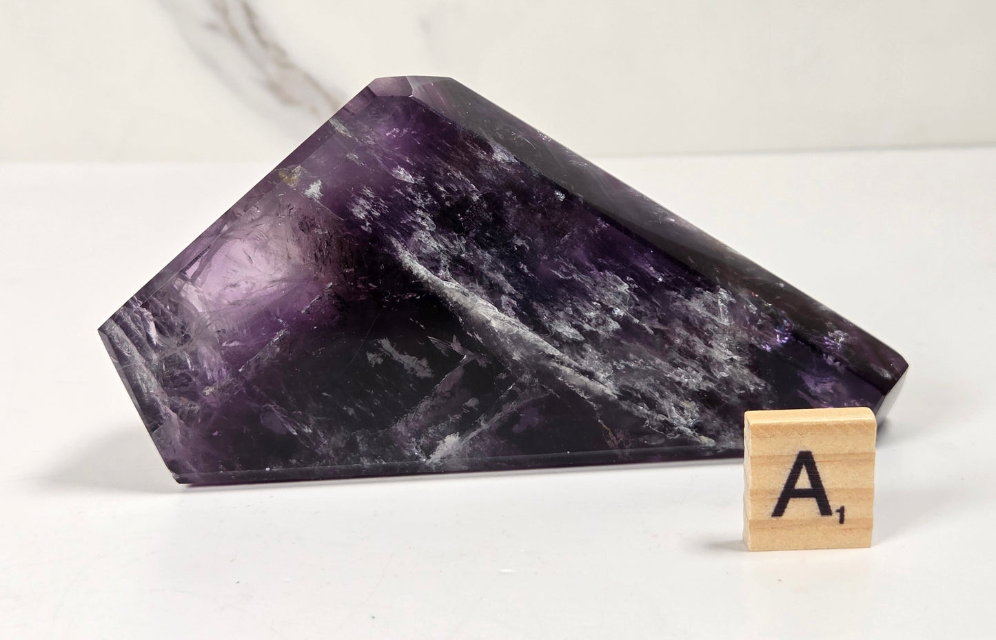 A polished purple amethyst crystal point displayed on a white surface next to a wooden Scrabble tile showing the letter A.
