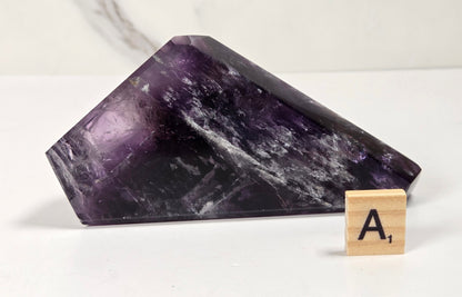 A polished purple amethyst crystal point displayed on a white surface next to a wooden Scrabble tile showing the letter A.