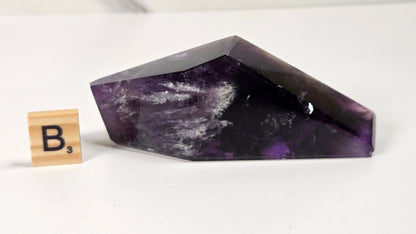A polished purple amethyst crystal point with natural white and dark inclusions, displayed on a white surface next to a small wooden Scrabble tile showing the letter B.