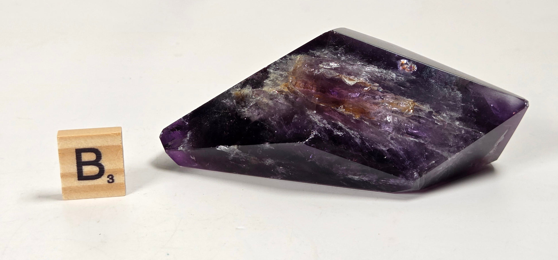 A polished purple amethyst crystal point with natural inclusions, displayed on a white surface next to a small wooden Scrabble tile showing the letter B.