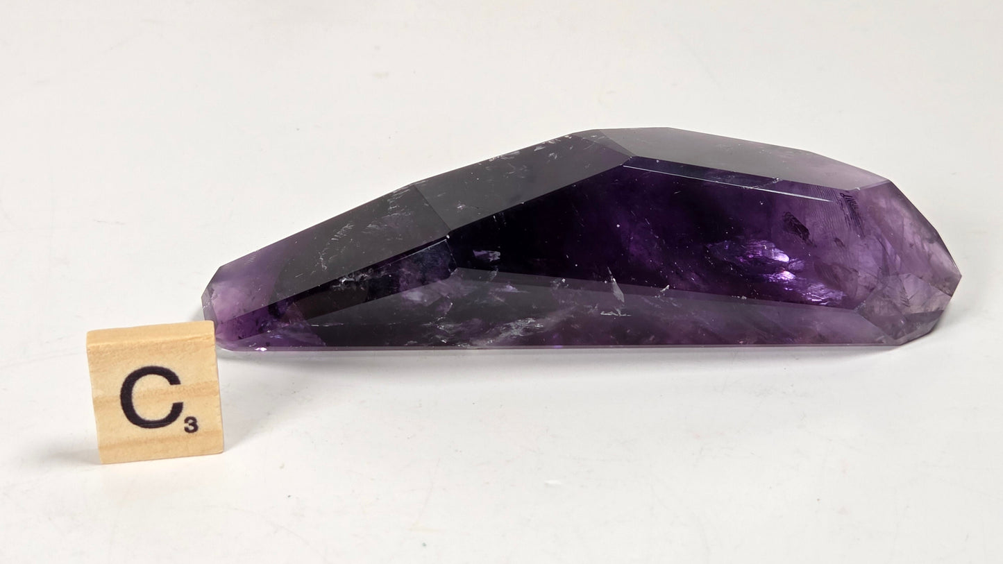A faceted purple amethyst crystal point shown on a white background next to a small wooden Scrabble tile marked C3.