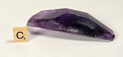 A faceted purple amethyst crystal point shown on a plain white background next to a small wooden Scrabble tile marked C3.