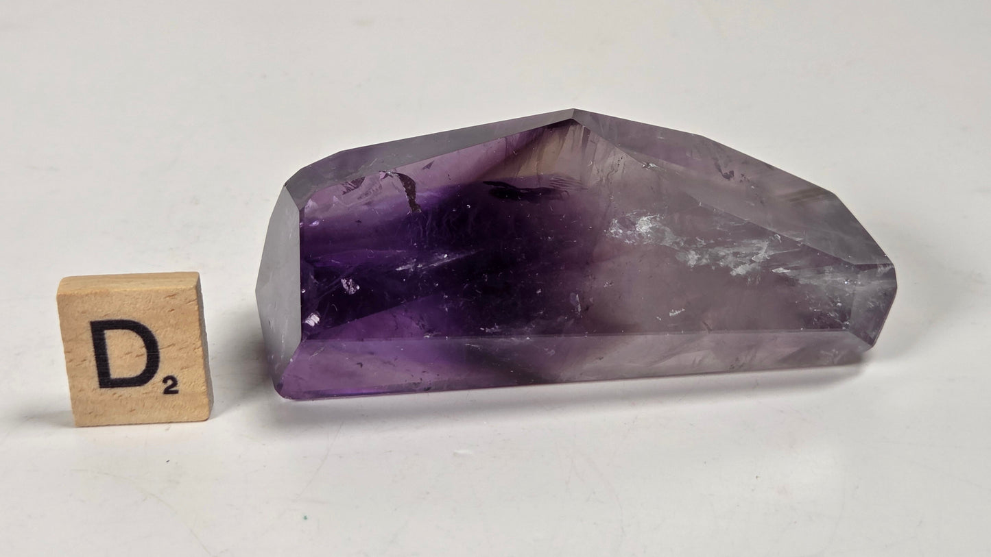 A faceted purple crystal point, likely amethyst, displayed on a white surface next to a wooden Scrabble tile showing the letter D.